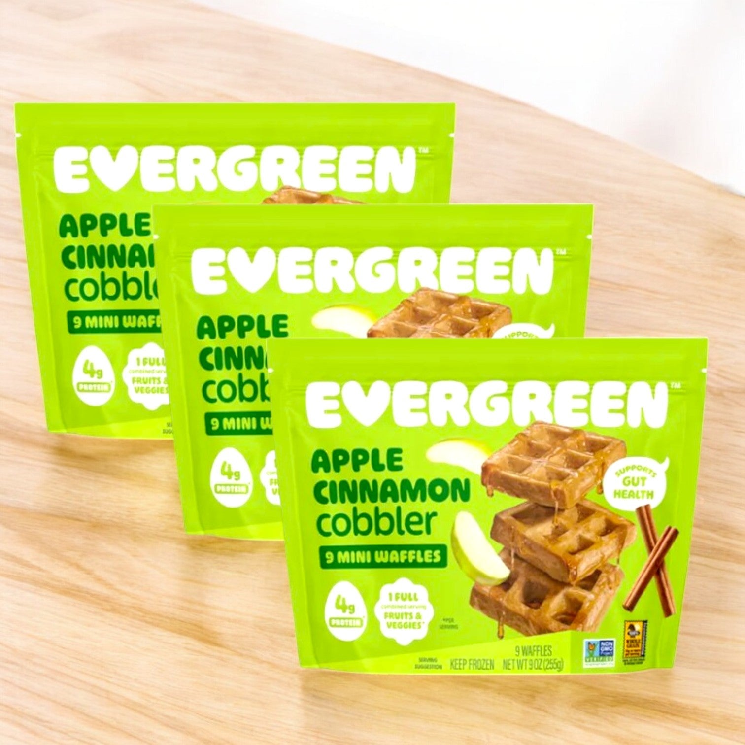 Three packs of Evergreen Apple Cinnamon Cobbler Frozen Mini Waffles (9 oz, 3 count) are displayed on a light wooden surface, ready to enjoy as a sweet snack.