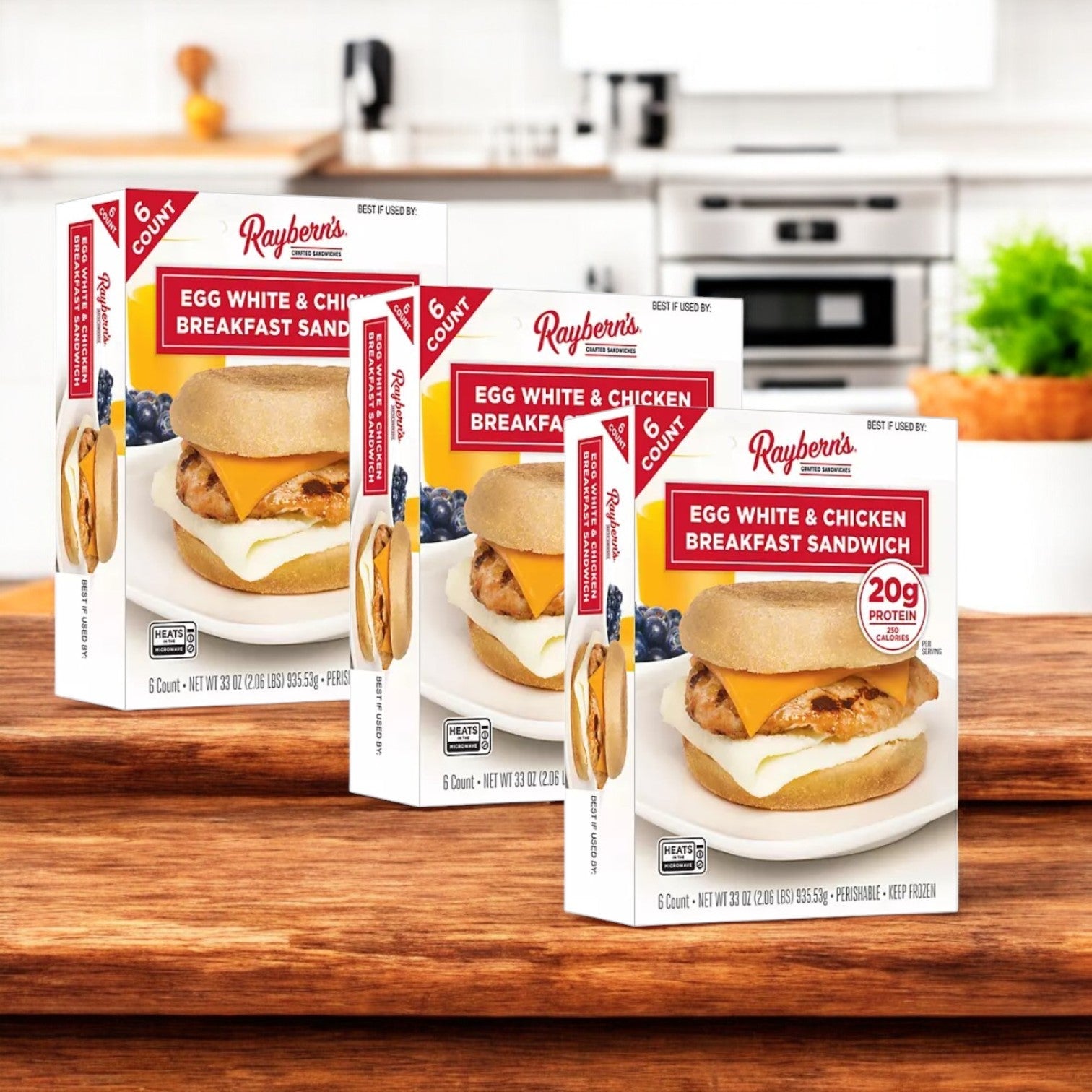 Three packs of Raybern's Egg White and Chicken Breakfast Sandwich are placed on a wooden surface in a kitchen setting, providing a protein-rich breakfast ideal for your low-carb routine.
