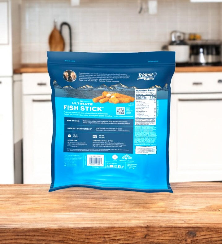 A 4 lbs. pack of Trident Seafoods Panko Breaded Wild Caught Alaska Pollock Fish Sticks rests on a wooden kitchen counter with the stove and utensils artistically blurred in the background.