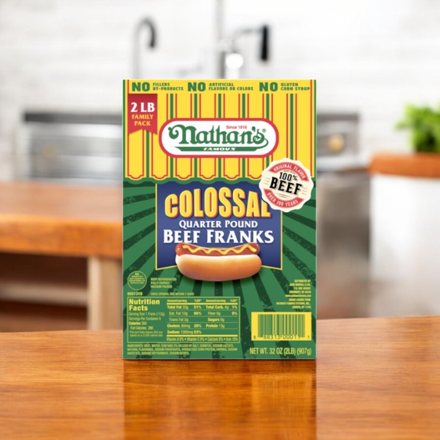 Nathan's Famous Beef Franks, Colossal Quarter Pound Family Pack-32 Oz-1 Pack, is displayed on a wooden table, featuring bold text and a hot dog image. The kitchen background with blurred plants and cutting board emphasizes these delights.