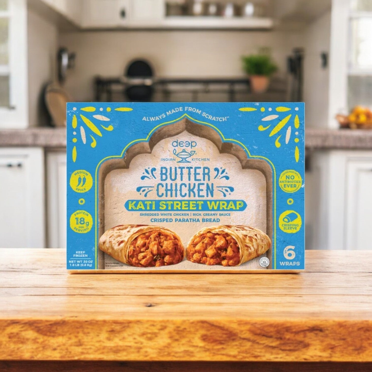 A case of Deep Indian Kitchen Kati Street Wrap, Butter Chicken (6 count) from Deep Indian Kitchen rests on a wooden kitchen counter with a blurred kitchen background.