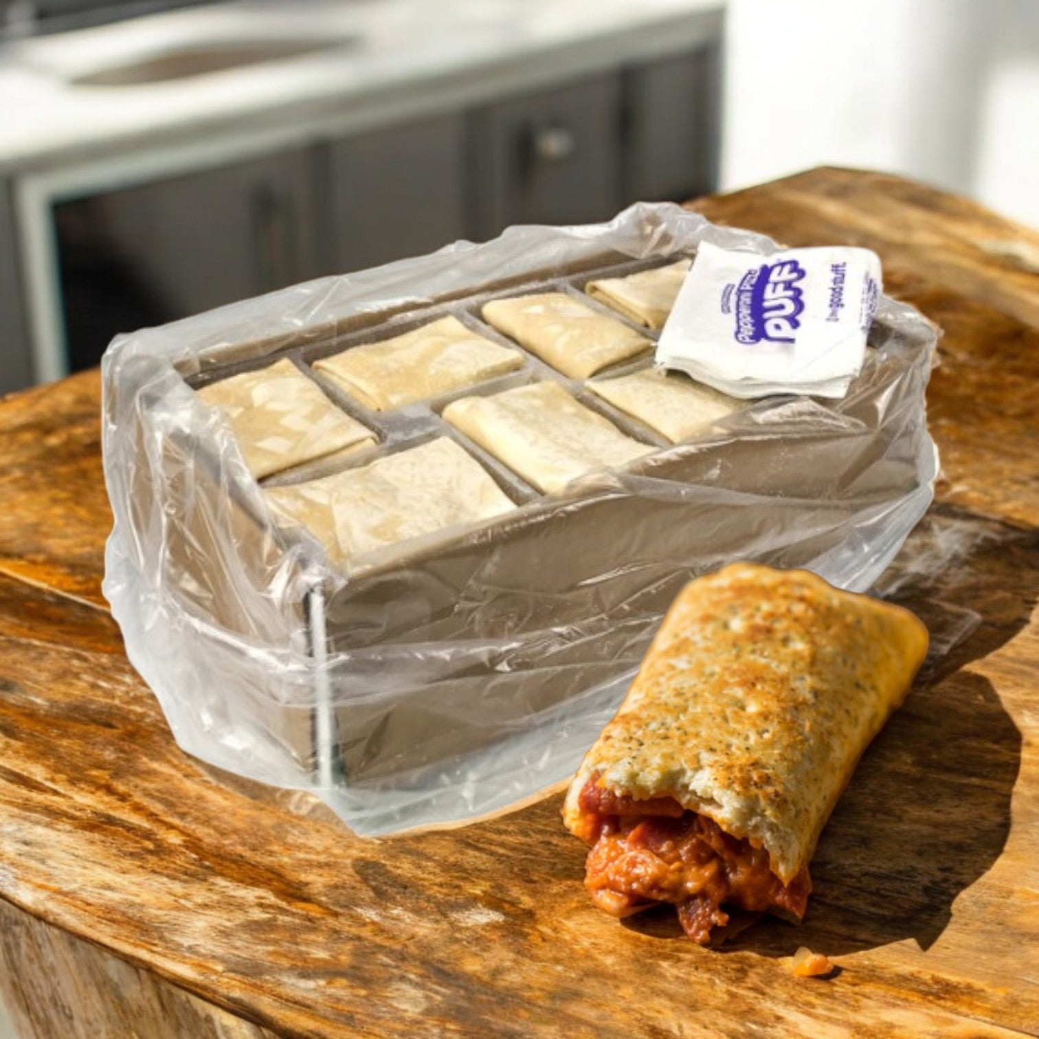 A case of Italco Puff Pepperoni Pizza (48 count, 6 oz each) by Itaco Foods in plastic packaging on a wooden surface, with one cooked puff cut open to reveal the filling.
