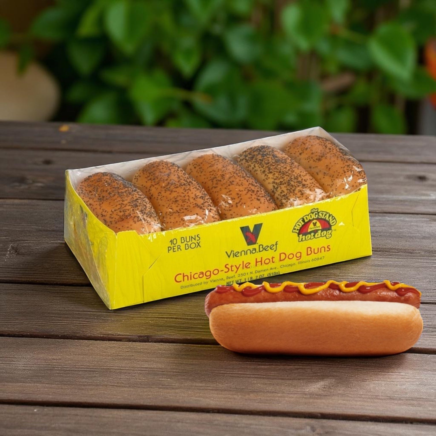 A 10-pack of Vienna Beef Vienna Poppy Seed Hot Dog Buns sits on a wooden table beside a hot dog topped with mustard and ketchup, with blurred green foliage in the background.