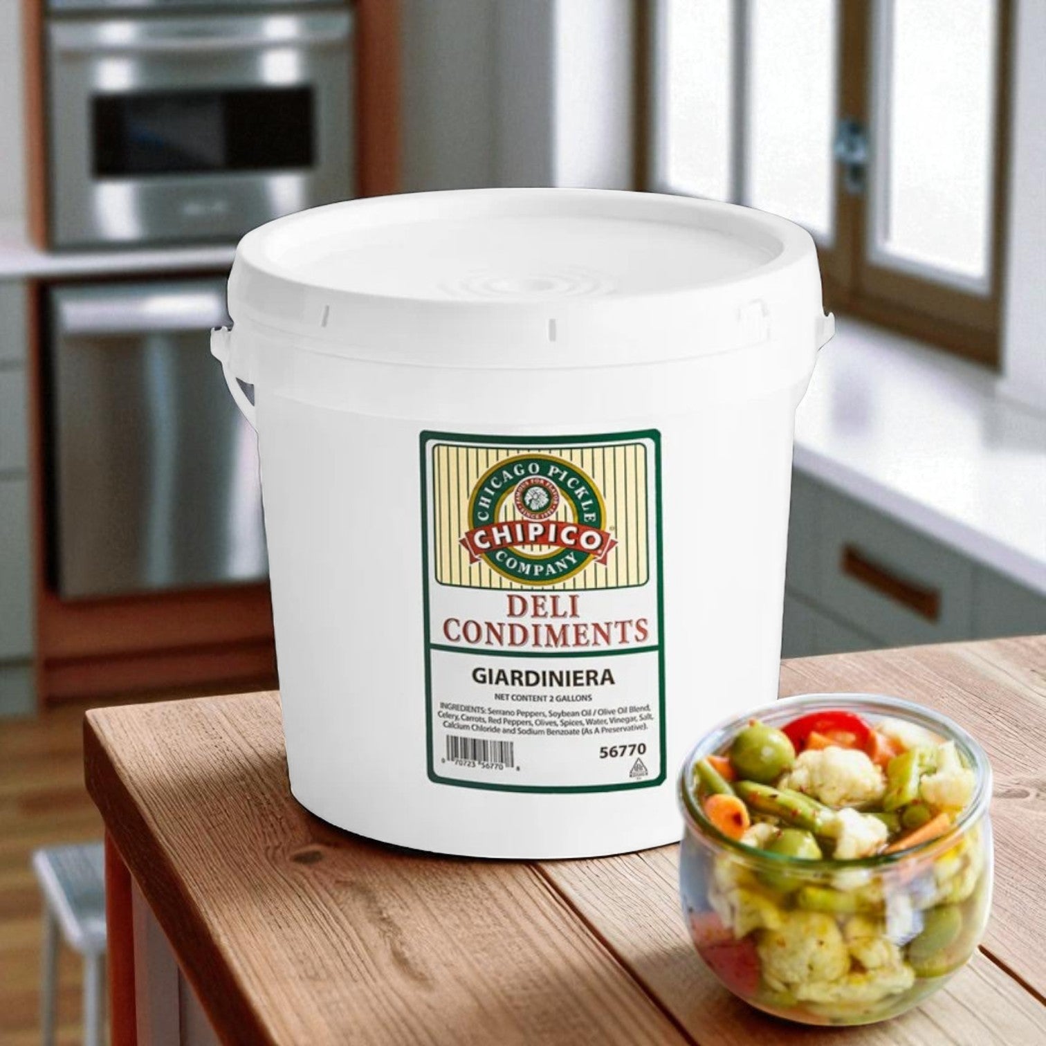 A white 2-gallon Vienna Beef Chipico Chicago Giardiniera container sits on a kitchen counter next to a glass bowl of mixed spicy relish and pickled vegetables.