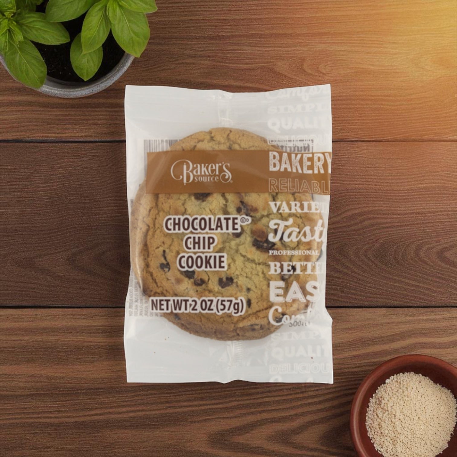 A Baker's Source Classic Cookie Chocolate Chip Individually Wrapped 2 Ounce sits on a wooden surface near a small green plant and a bowl of light brown granules, ideal for anyone needing bulk cookies—48 count per box—for any occasion.