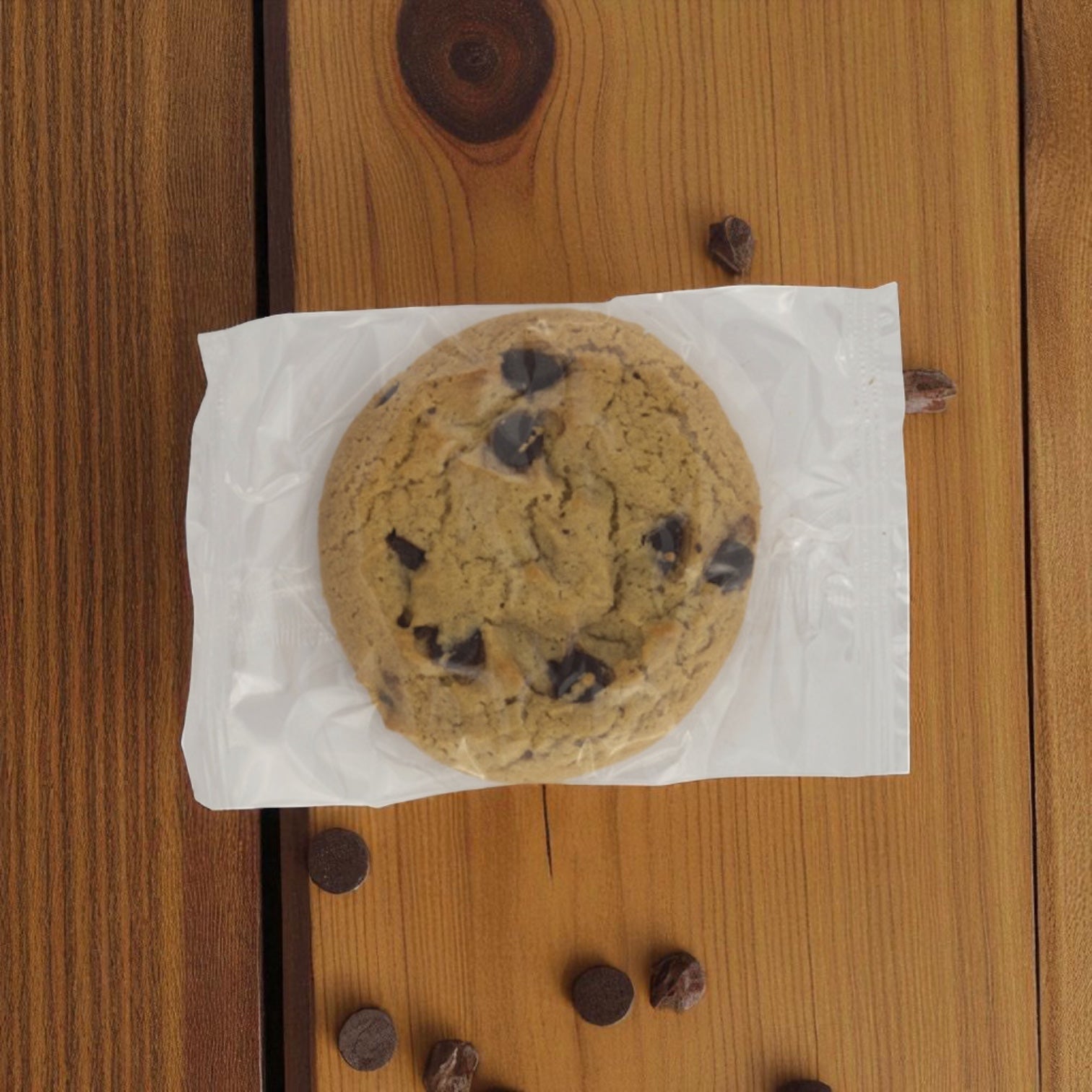 A Baker's Source Classic Cookie Chocolate Chip Clear Individually Wrapped 1 oz sits on a wooden surface, with scattered chocolate chips nearby. Packaged for freshness, available in a 144-count box.