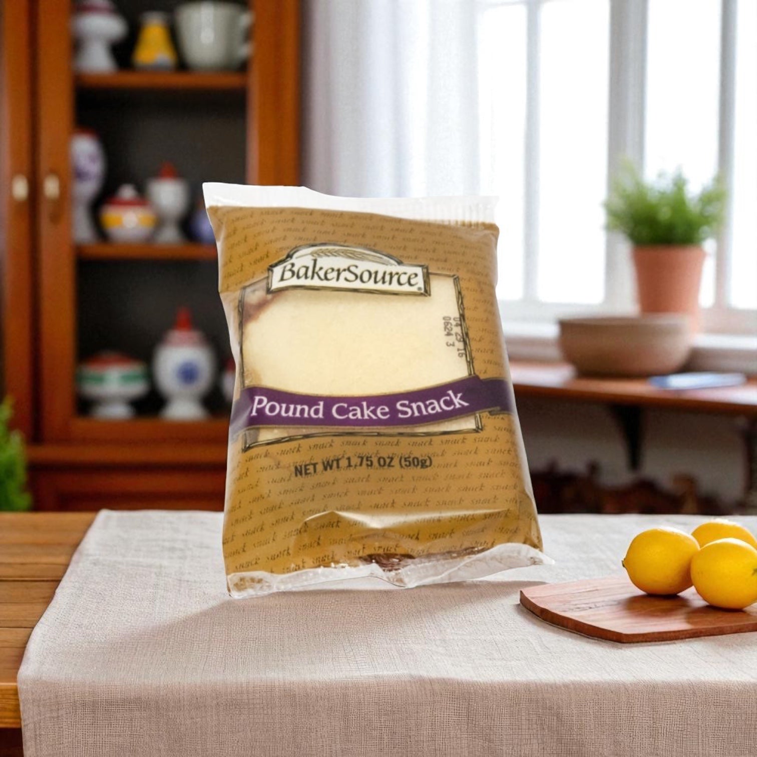 A Baker's Source Classic Pound Cake, individually wrapped (1.75 oz, 24 count), sits on a cloth-covered table with lemons and a wooden cabinet in the background—ideal for grab-and-go convenience.