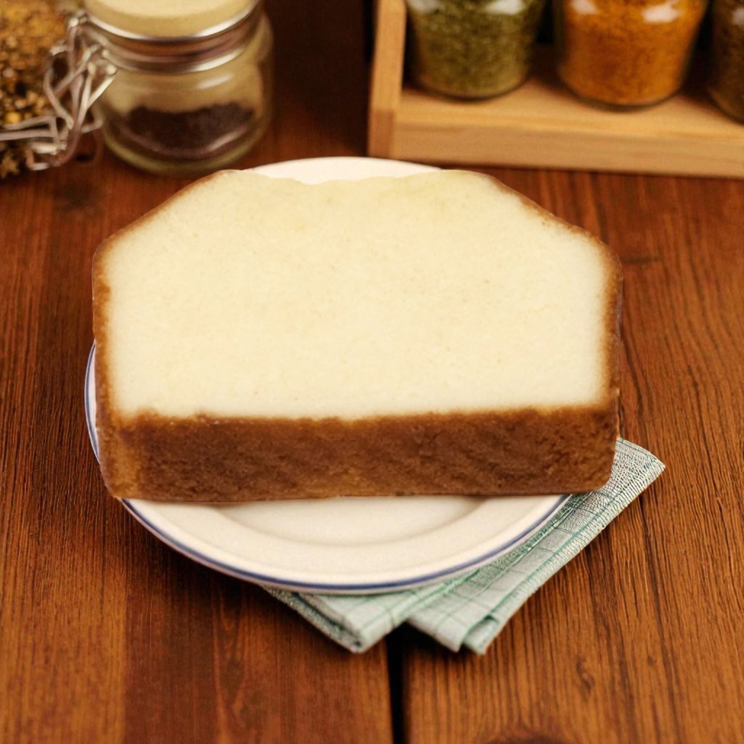 A slice of plain white bread sits on a plate with a green napkin and spice jars in the background—ideal for a quick snack or pairing with Baker's Source Classic Individually Wrapped Pound Cake, 1.75 oz (24 count).