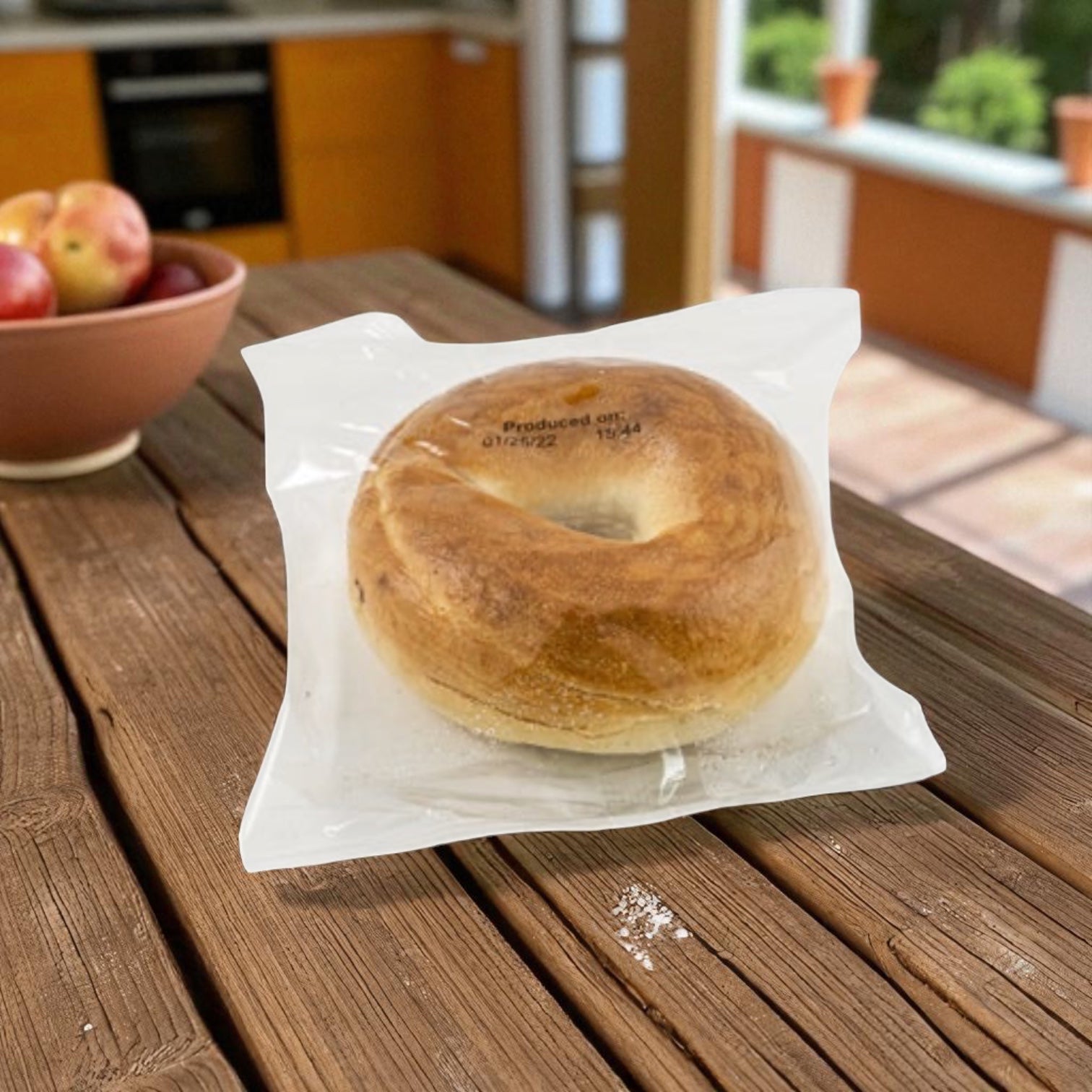 A Baker's Source Classic Bagel Plain Individually Wrapped Sliced 3 oz sits on a kitchen table with apples and a window in the background.