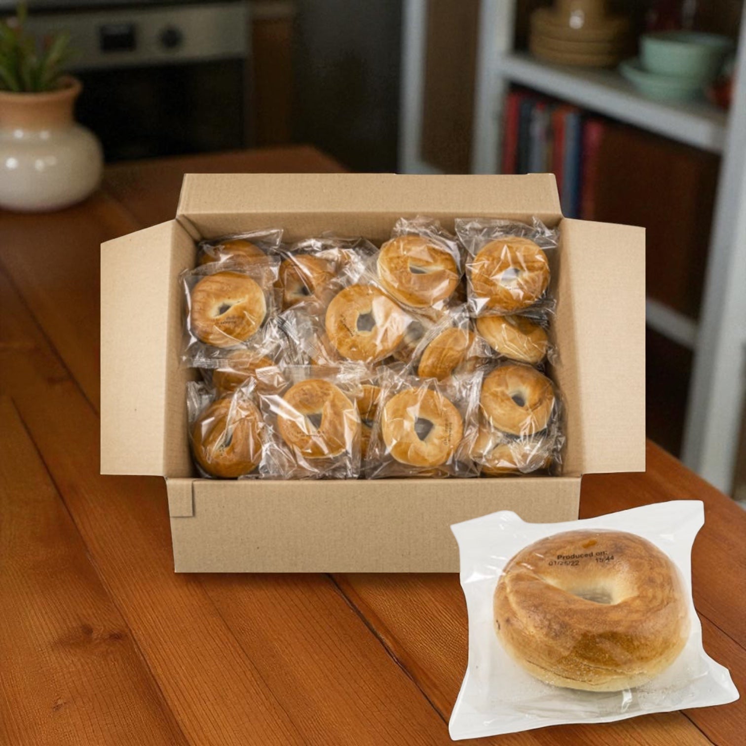 A box of Baker's Source Classic Bagel Plain Individually Wrapped Sliced 3 oz - 60 Count sits on a wooden table, with each bagel individually wrapped and one vacuum-sealed bagel displayed in front.