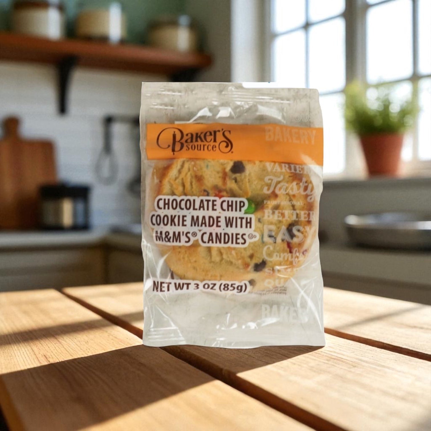 A box of Baker's Source Classic Cookie Chocolate Chip With Candy, 3 oz individually wrapped (48 count), sits on a wooden table in a sunlit kitchen, featuring delicious cookies perfect for snacking anytime.