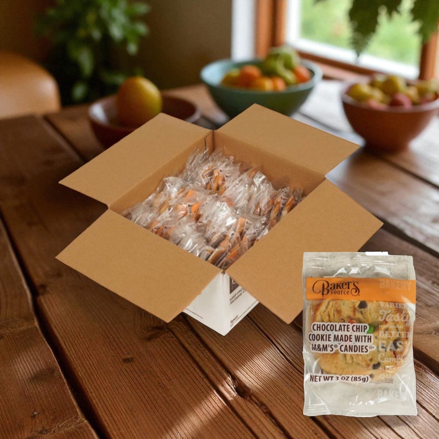 A box of Baker's Source Classic Cookie Chocolate Chip With Candy Individually Wrapped 3 oz - 48 Count sits on a wooden table with fruit bowls behind it. An enlarged packet of the cookie is displayed in the foreground.