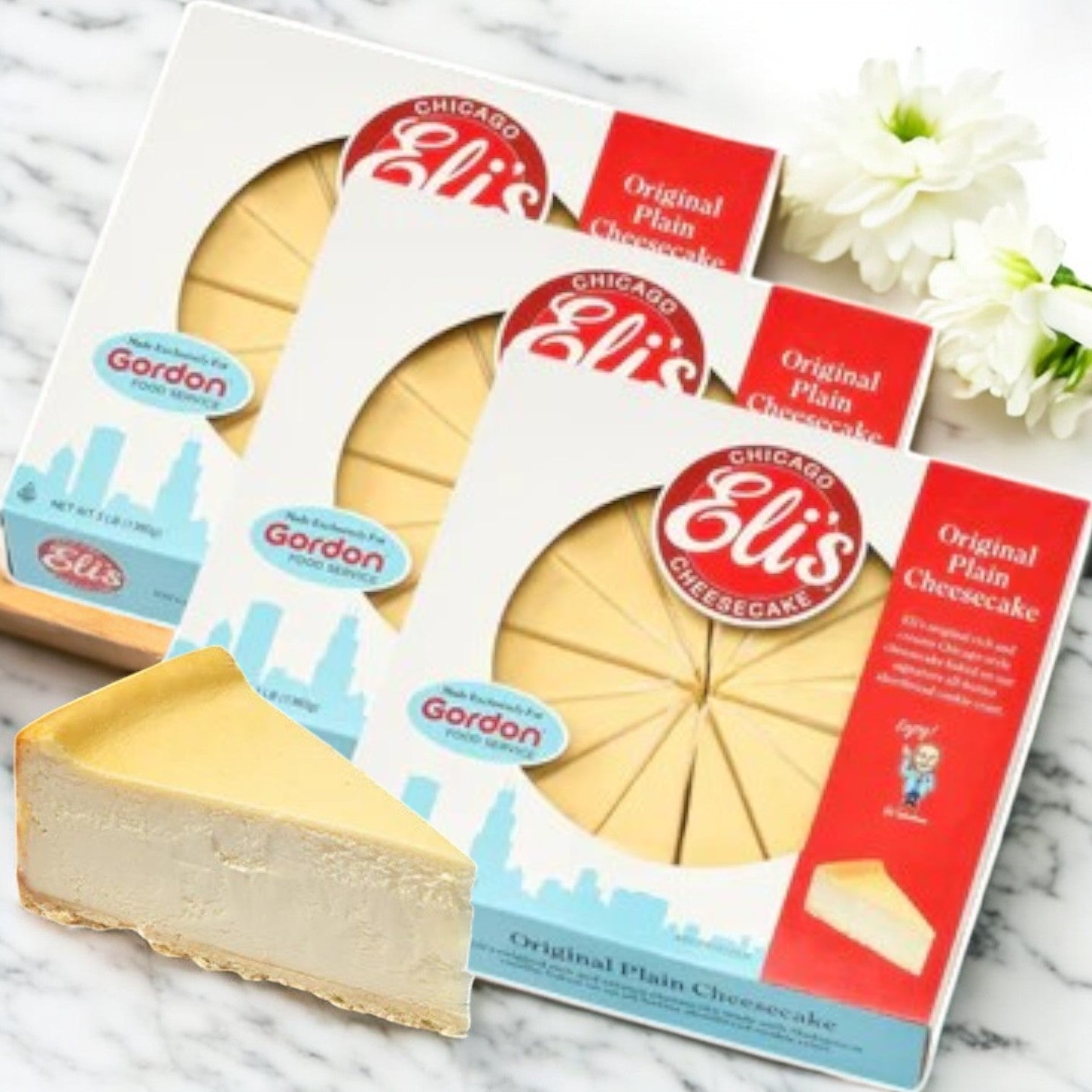 Eli's 9" Original Plain Cheesecake- 3 Count boxes are elegantly arranged on a marble surface, with an extra slice featuring its buttery shortbread cookie crust prominently displayed. Delicate white flowers enhance the background, highlighting the renowned taste of Eli's Cheesecake.