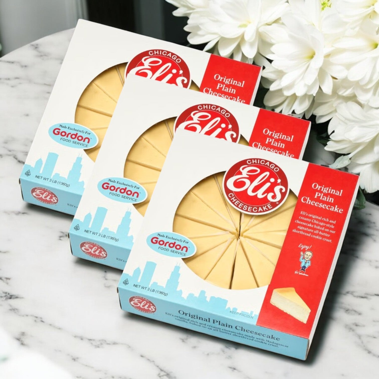 Three Eli's 9" Original Plain Cheesecakes, each featuring a buttery shortbread cookie crust, are elegantly displayed on a marble surface with delicate white flowers in the background.