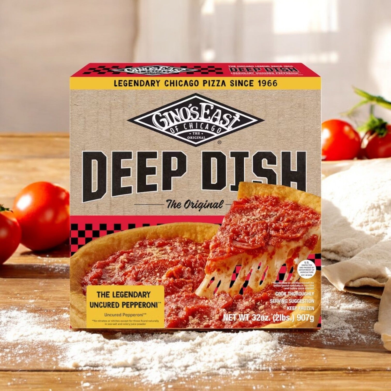 A Gino's East Pizza box rests on a wooden surface, showcasing its buttery crust. The Uncured Pepperoni Deep Dish pizza is adorned with pepperoni, while a powdered table and ripe tomatoes enhance the appealing scene.