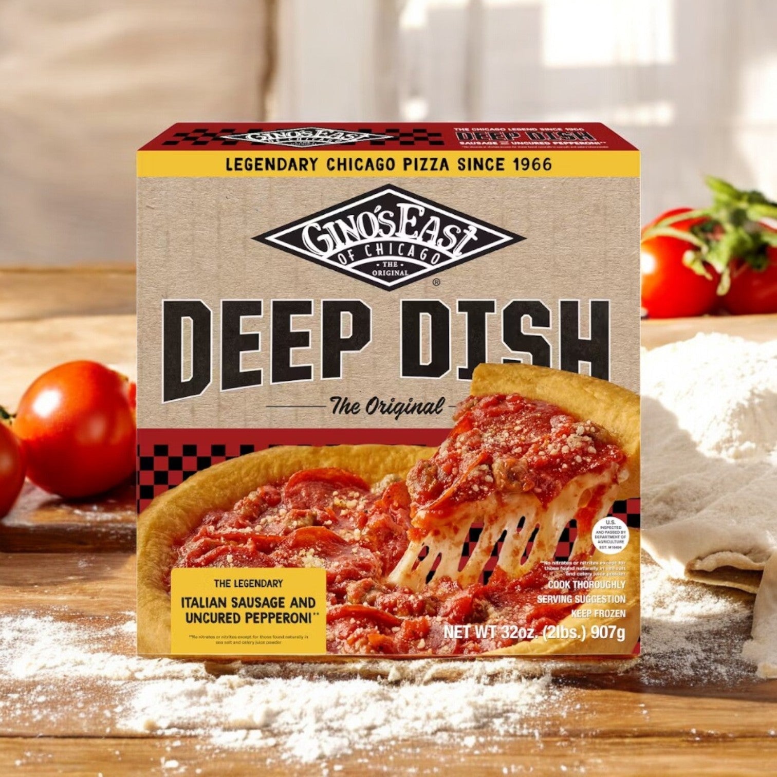 A box of Gino's East Pizza, Sausage & Uncured Pepperoni, Crumbled, Italian Deep Dish 32 oz., sits on a flour-dusted surface with fresh tomatoes and greenery.