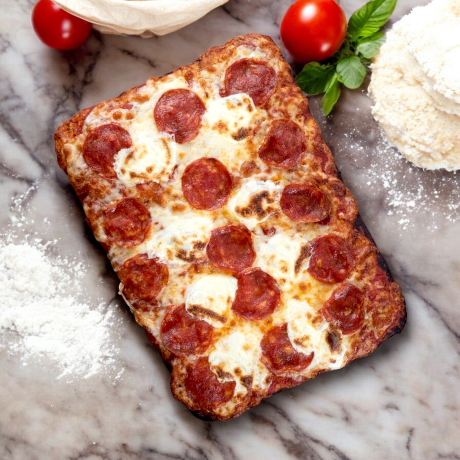 A Massa Pizza Company Pepperoni Pizza – 21.5 oz. sits on a marble surface, exuding craftsmanship with scattered flour, whole tomatoes, basil leaves, and stacks of premium grated cheese nearby.