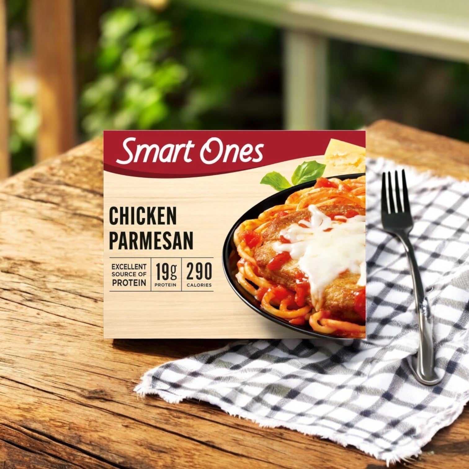A box of Smart Ones Chicken Parmesan with Spaghetti, Marinara Sauce & Mozzarella Cheese rests on a checkered cloth outdoors. The package notes 19g protein and 290 calories, a fork beside it completes the scene.