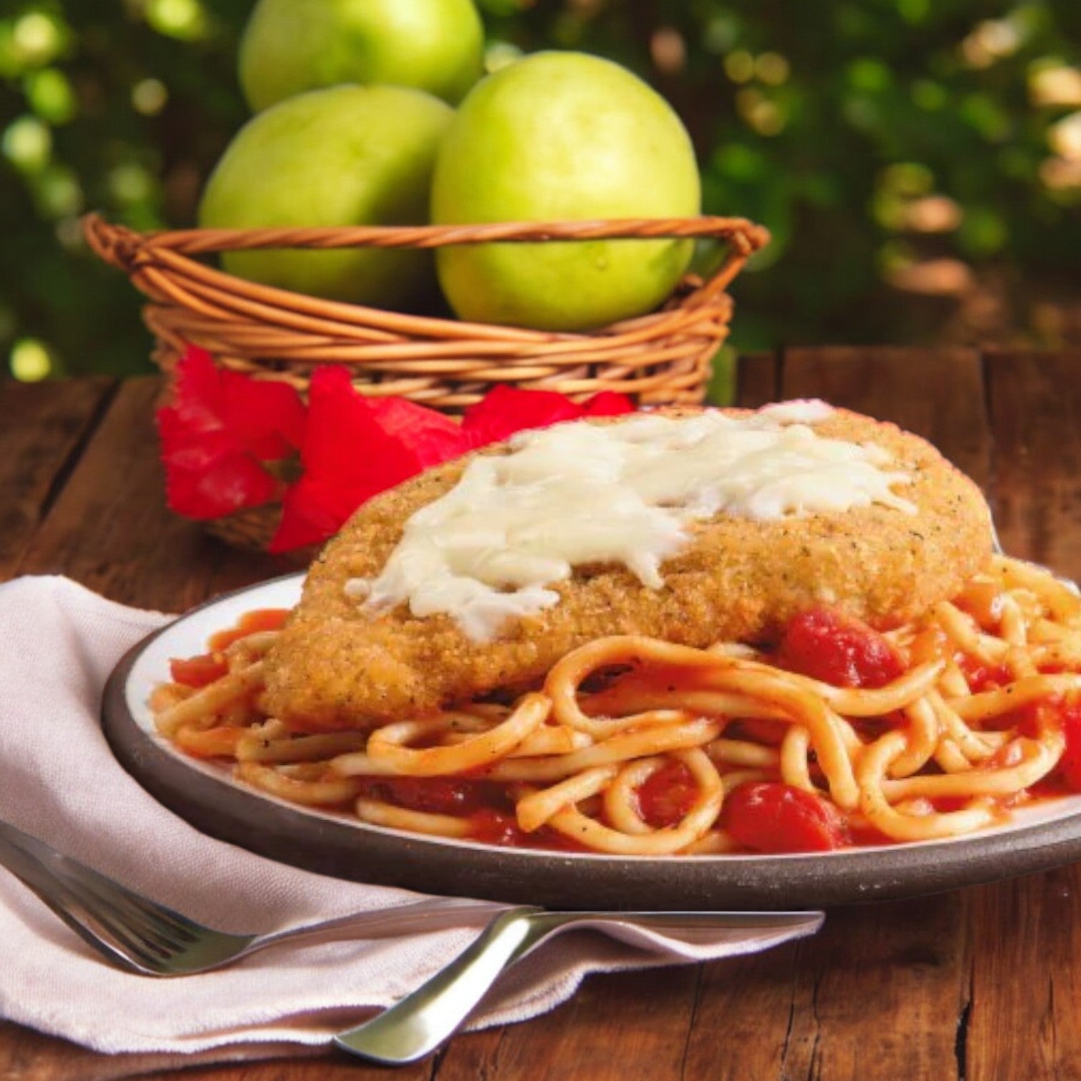 A plate of Smart Ones Chicken Parmesan with Spaghetti, Marinara Sauce & Mozzarella Cheese Frozen Meal is set on a wooden table. A fork and napkin lie beside it, while a basket of green apples in the background adds freshness to this delightful meal.
