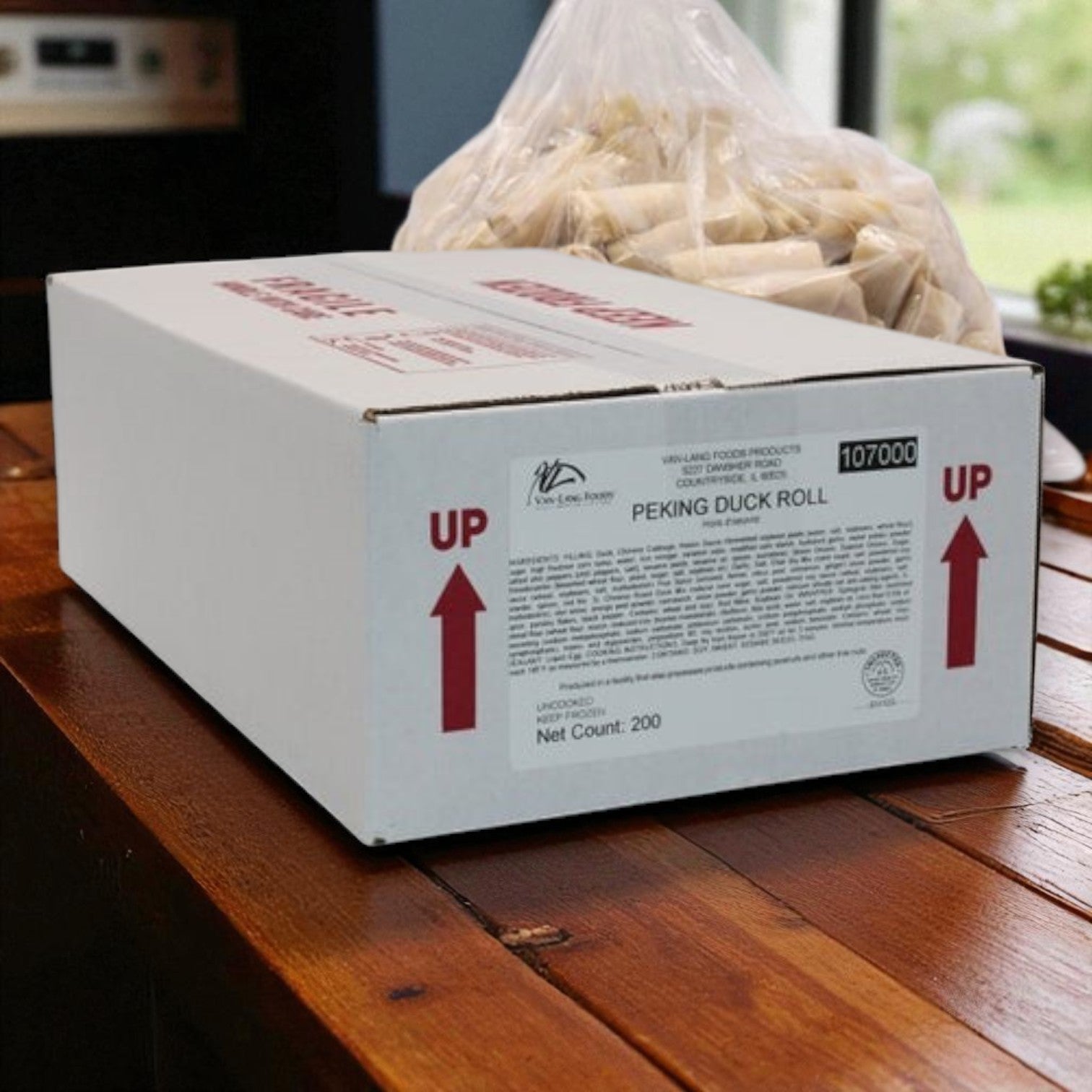 A white box labeled "Van Lang Peking Duck Spring Rolls – 200 Count (Fully Cooked, Frozen)" sits on a wooden table; ideal for catering, these gourmet appetizers promise authentic flavor. A bag of food items is visible in the background.