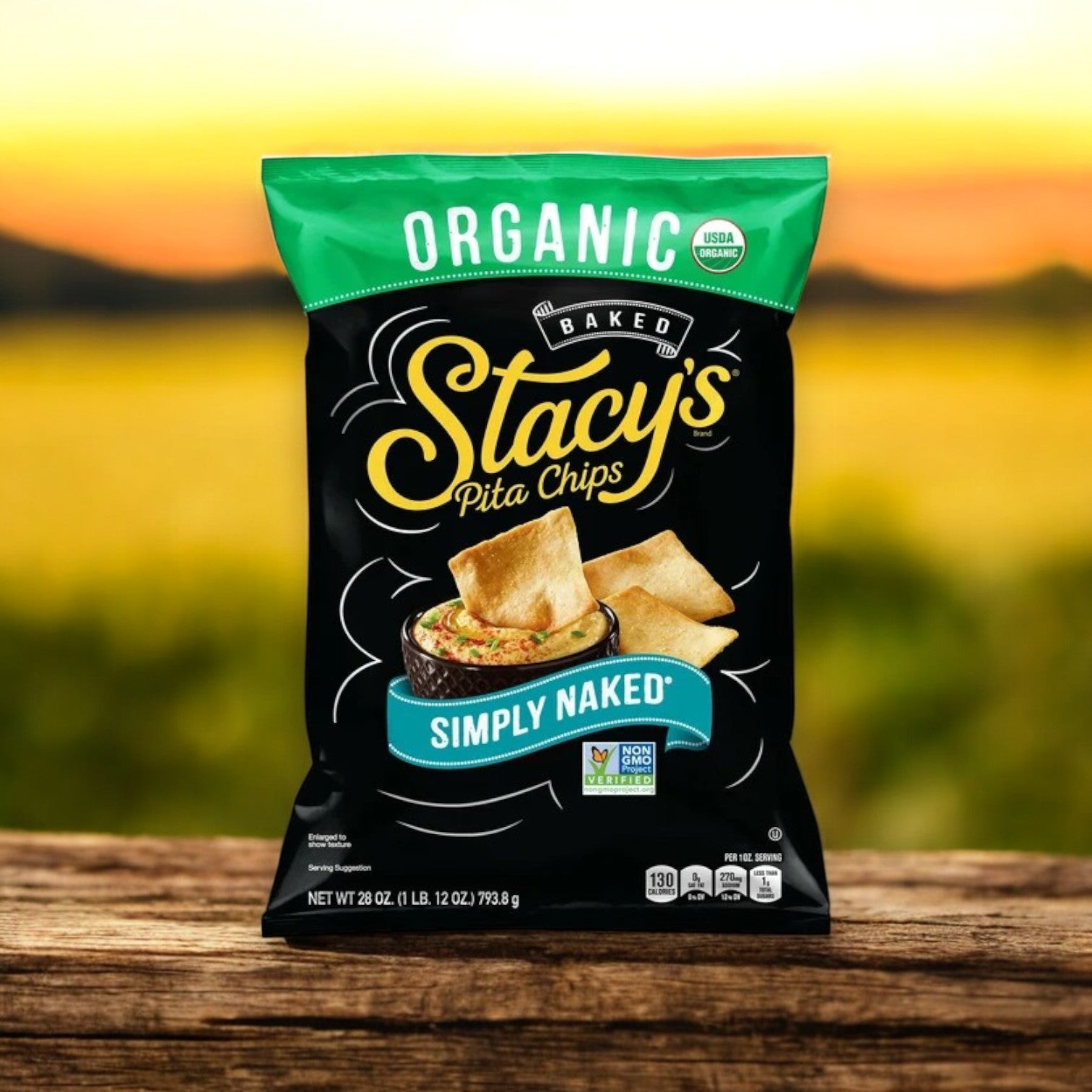A 28 oz. bag of Stacy's Organic Simply Naked Pita Chips is elegantly placed on a wooden surface against a blurred outdoor background, showcasing it as the perfect sea salt snack.