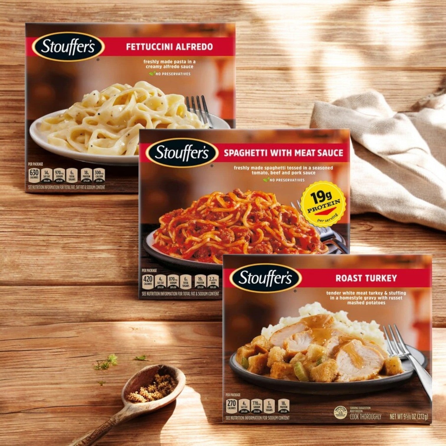 Three Stouffer's entrées, Fettuccini Alfredo, Spaghetti with Meat Sauce, and Roast Turkey from the Stouffer's Bundle! 3 Total!, sit on a wooden surface. Sunlight highlights them alongside a small bowl of seasoning, ideal for enhancing these comforting meals.
