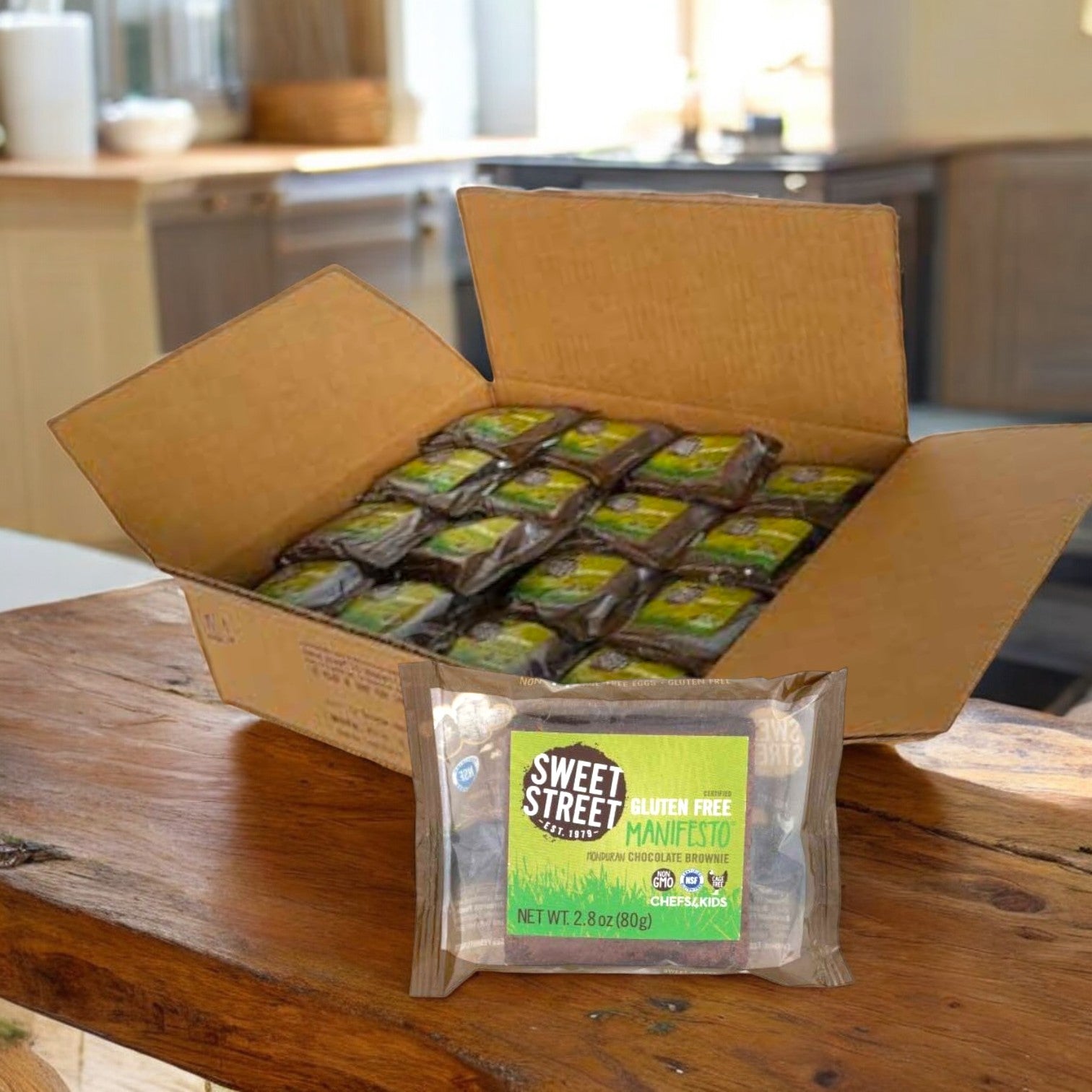 An open cardboard box on a wooden table holds Sweet Street's individually wrapped 2.8 oz. Honduran Chocolate Manifesto Brownies in clear packaging, crafted with rich chocolate for a tempting, gluten-free dessert menu addition; available 48 per case.