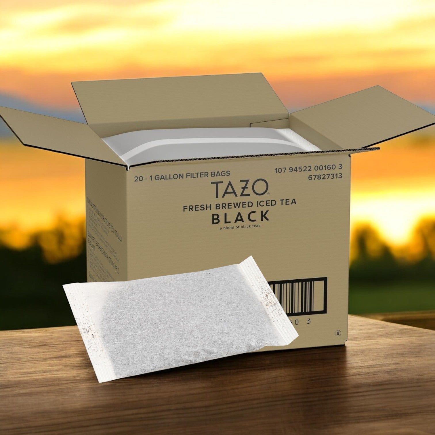 An open Tazo Tea Iced Black - 20/1 Gallon box rests on a wooden surface, showcasing a large filter bag ready for brewing. In the backdrop, a sunset glow bathes the blurred landscape, creating a perfect setting to enjoy this bold and refreshing iced tea.