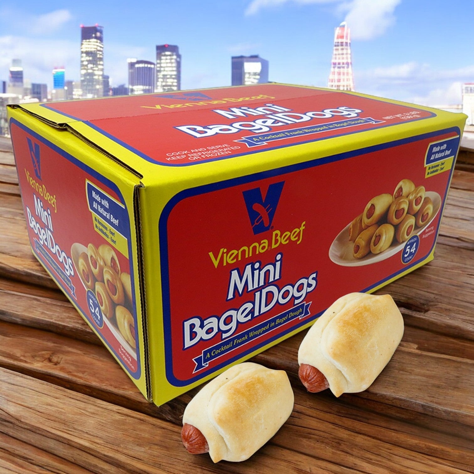 A box of Vienna Beef Mini Bagel Dogs (54 count) sits on a wooden surface with two in front, against a picturesque city skyline.