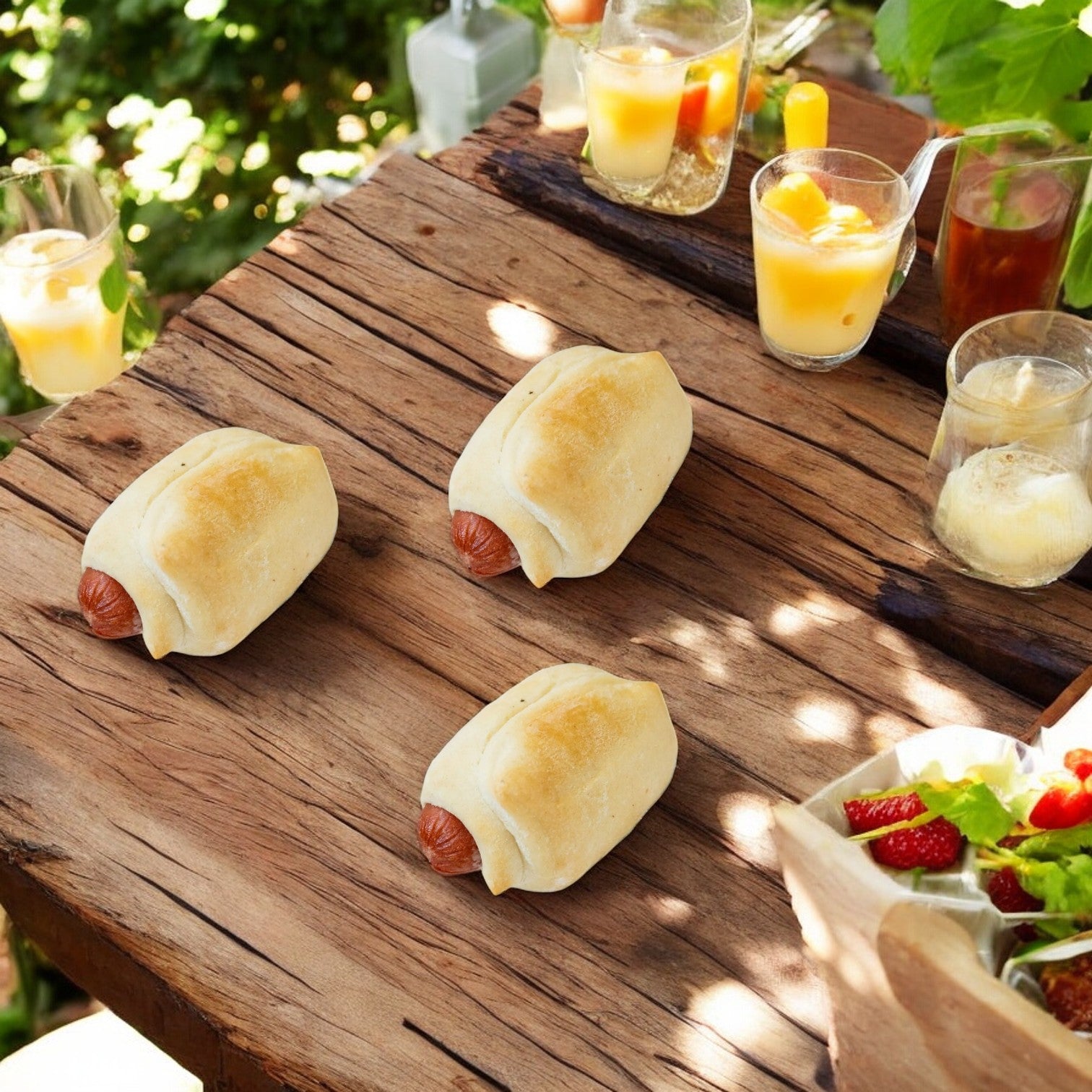 Three Vienna Beef Mini Bagel Dogs rest on a rustic wooden table, surrounded by refreshing drinks and a vibrant fruit salad, creating the perfect outdoor picnic spread.