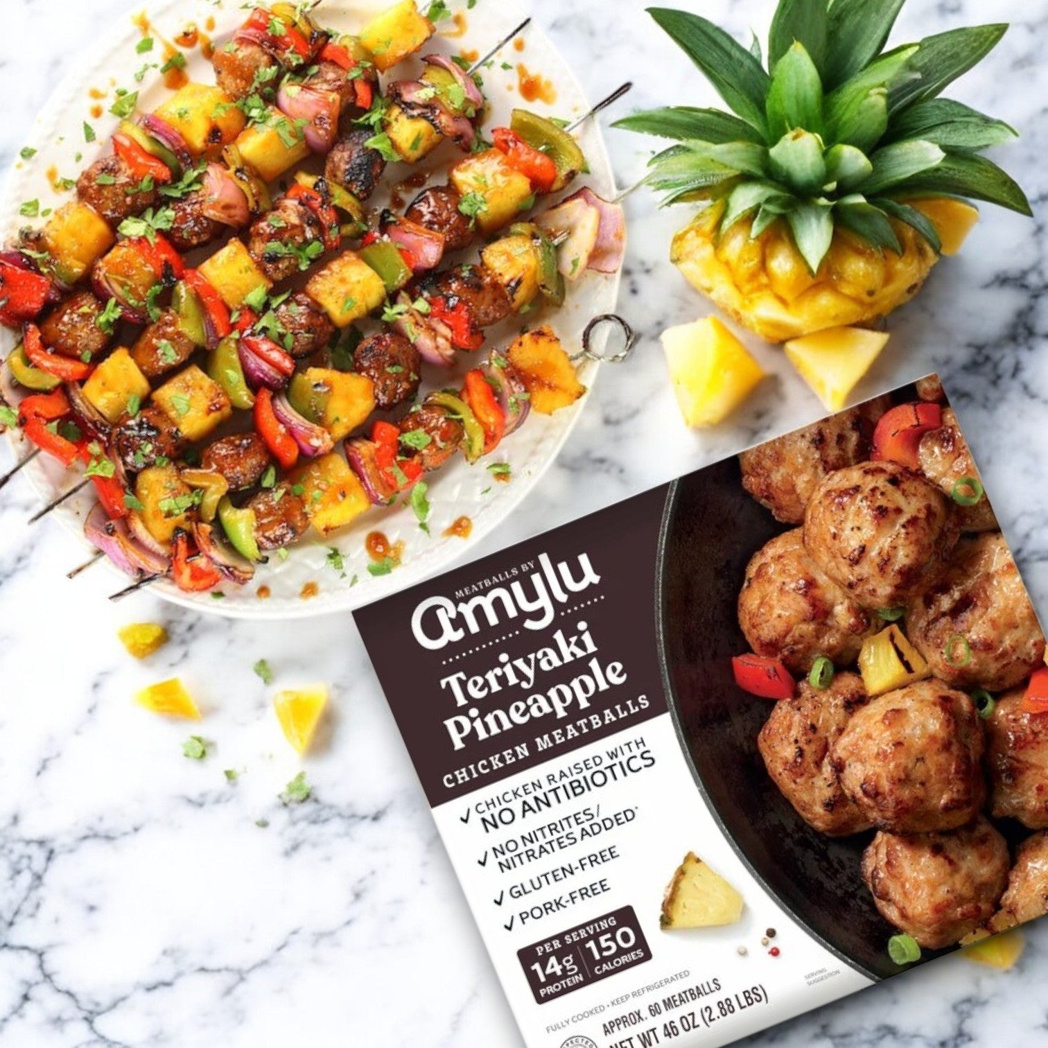 Skewered teriyaki pineapple chicken and vegetables grace a marble surface alongside a whole pineapple and Amylu's Teriyaki Pineapple Chicken Meatballs, 46 oz in 3 packs. Made with antibiotic-free chicken, these meatballs perfectly complement the teriyaki-glazed dish.