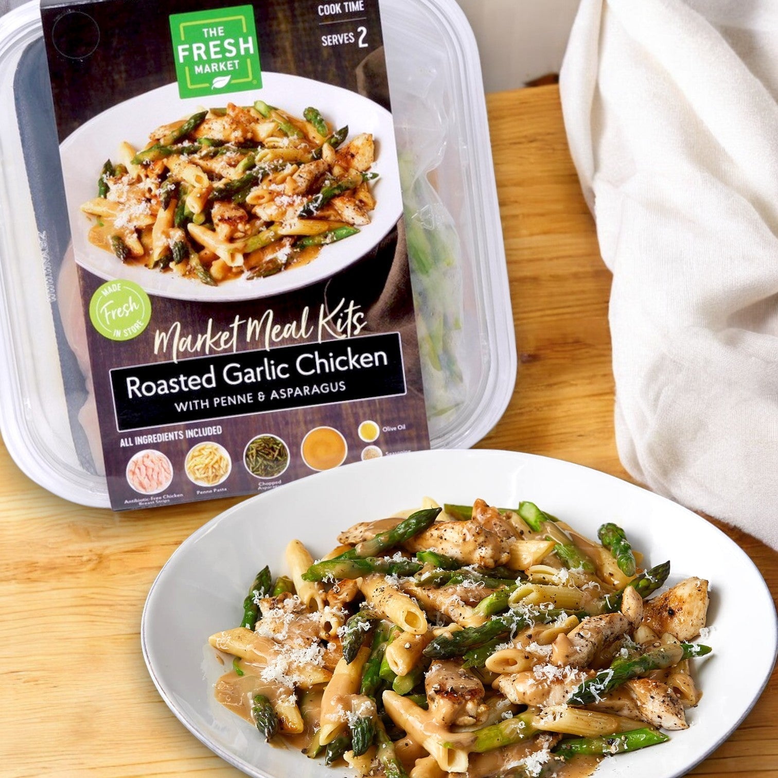 A meal kit labeled "The Fresh Market Roasted Garlic Chicken with Penne & Asparagus Market Meal Kit – 36 oz" sits next to a plated serving on a wooden surface.