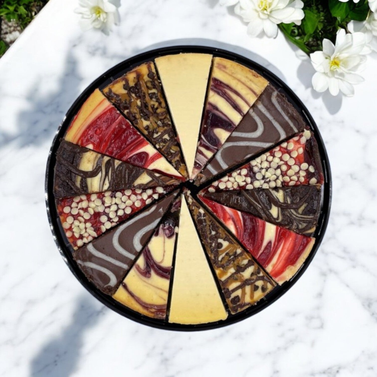 The Father's Table Variety Cheesecake, featuring premium ingredients and a selection of flavors and toppings, is elegantly sliced into 14 pieces. Displayed on a marble surface beside white flowers, the 10-inch cheesecake from The Father's Table highlights its exquisite quality.