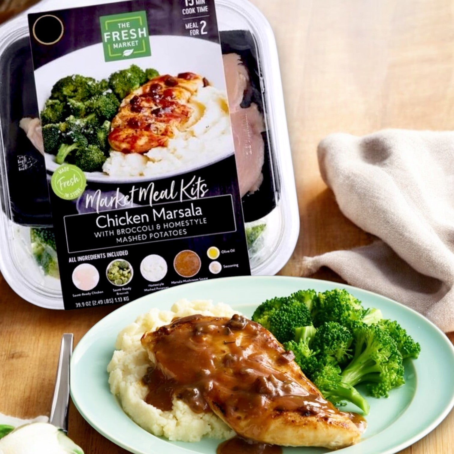 The Fresh Market Chicken Marsala with Broccoli & Homestyle Mashed Potatoes Market Meal Kit – 39 oz. sits elegantly beside a plated serving on a light green plate, complete with mashed potatoes and broccoli, on a wooden table adorned with a beige cloth.