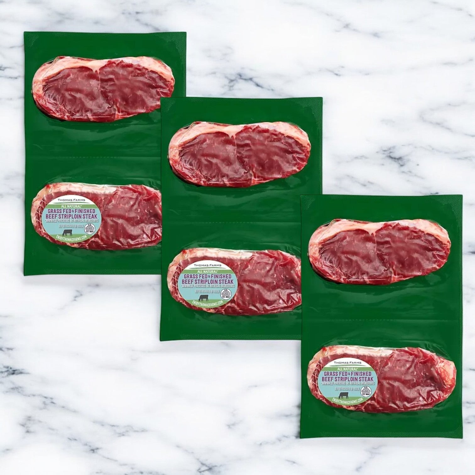 Three vacuum-sealed packages of premium Thomas Farms Grass Fed New York Strip Steaks are elegantly displayed on a marble surface.