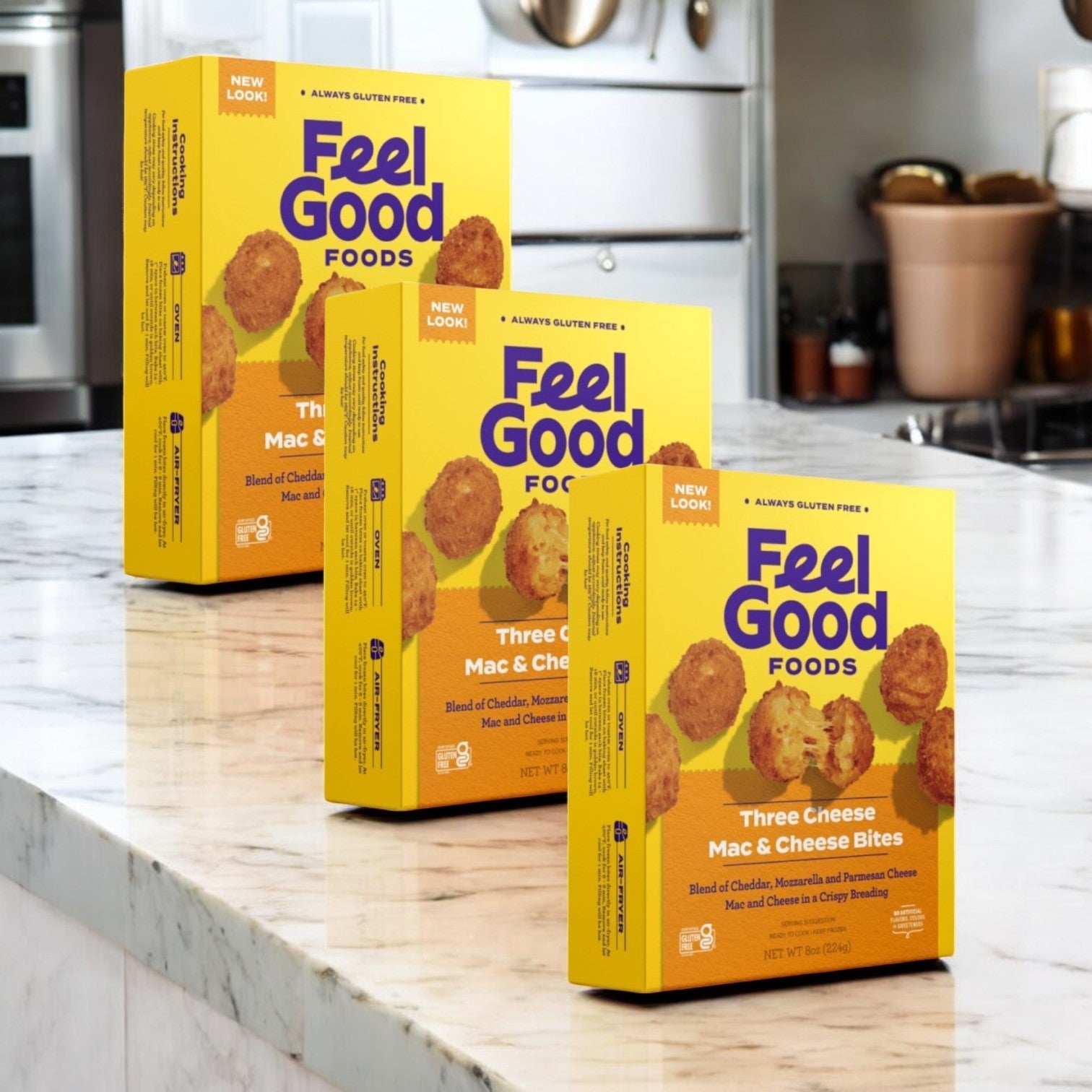 Three boxes of Feel Good Foods Three Cheese Mac & Cheese Bites (8 oz. each) sit on a marble counter. Utensils and decor are visible, making these gluten-free bites the ideal snack appetizer to impress guests.