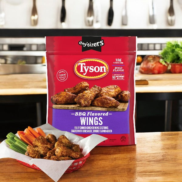 A Tyson Any'tizers BBQ Flavored Chicken Wings package is on the kitchen counter, alongside a basket of cooked BBQ wings and fresh vegetables, creating the ideal ready-to-cook snack.
