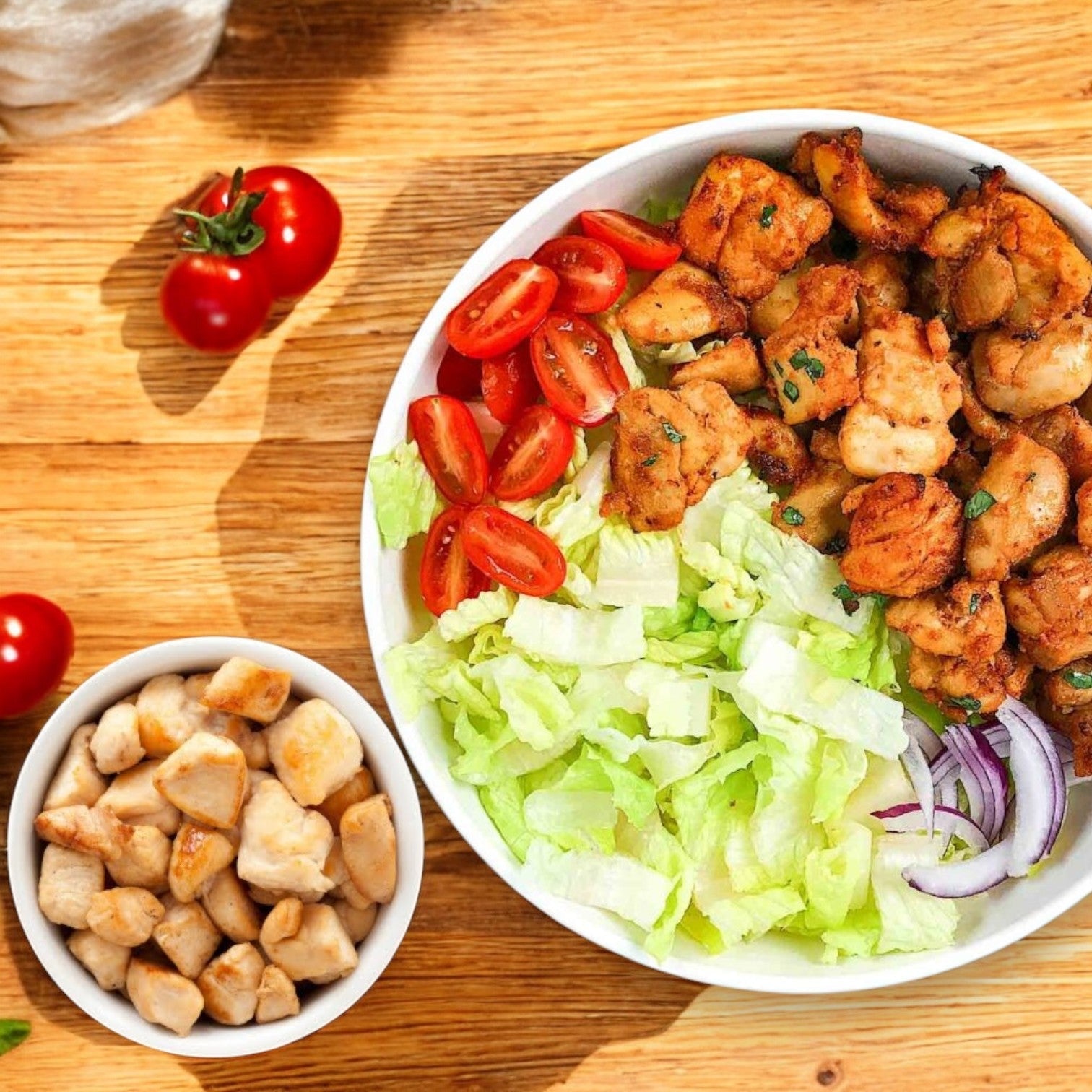 A bowl on a wooden table features fully cooked all-natural Tyson Red Label Diced Chicken Breast, sliced cherry tomatoes, lettuce, and red onion. Nearby, a small bowl with extra diced chicken breast and whole cherry tomatoes completes the fresh setup.