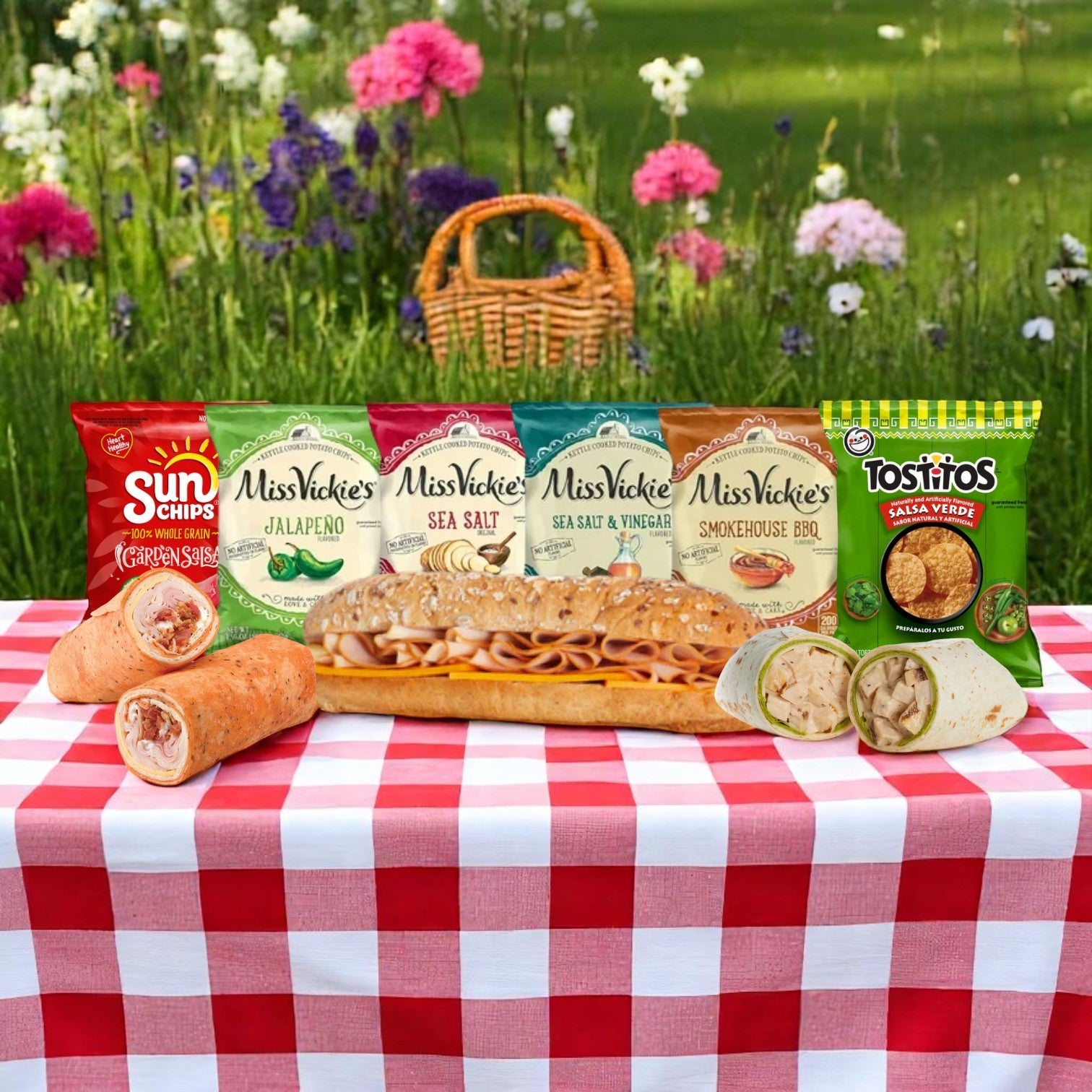 A picnic table shows the E.A. Sween 10-Pack Wrap & Sub Sandwich Bundle with 10 assorted snack chips on a red and white checkered cloth in a garden, surrounded by flowers and a wicker basket.