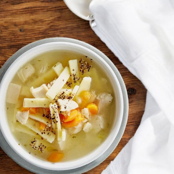 A bowl of Bistro Soups by Vienna Beef – Mama's Chicken Noodle Soup sits on a wooden table beside a white cloth, exuding warmth and comfort, akin to enjoying Vienna Beef Hot Dog Buns fresh from the oven.