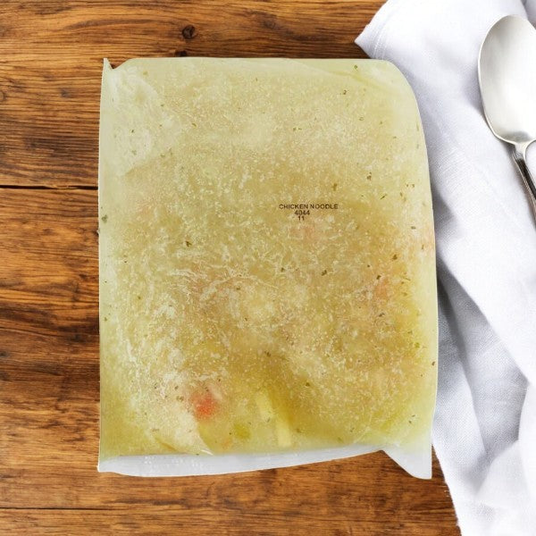 A case of Bistro Soups by Vienna Beef—Mama's Chicken Noodle Soup, 4 lb., sits sealed on a wooden surface with a folded white cloth and spoon, while nearby frozen poppy seed-topped hot dog buns add an unexpected twist to this cozy scene.