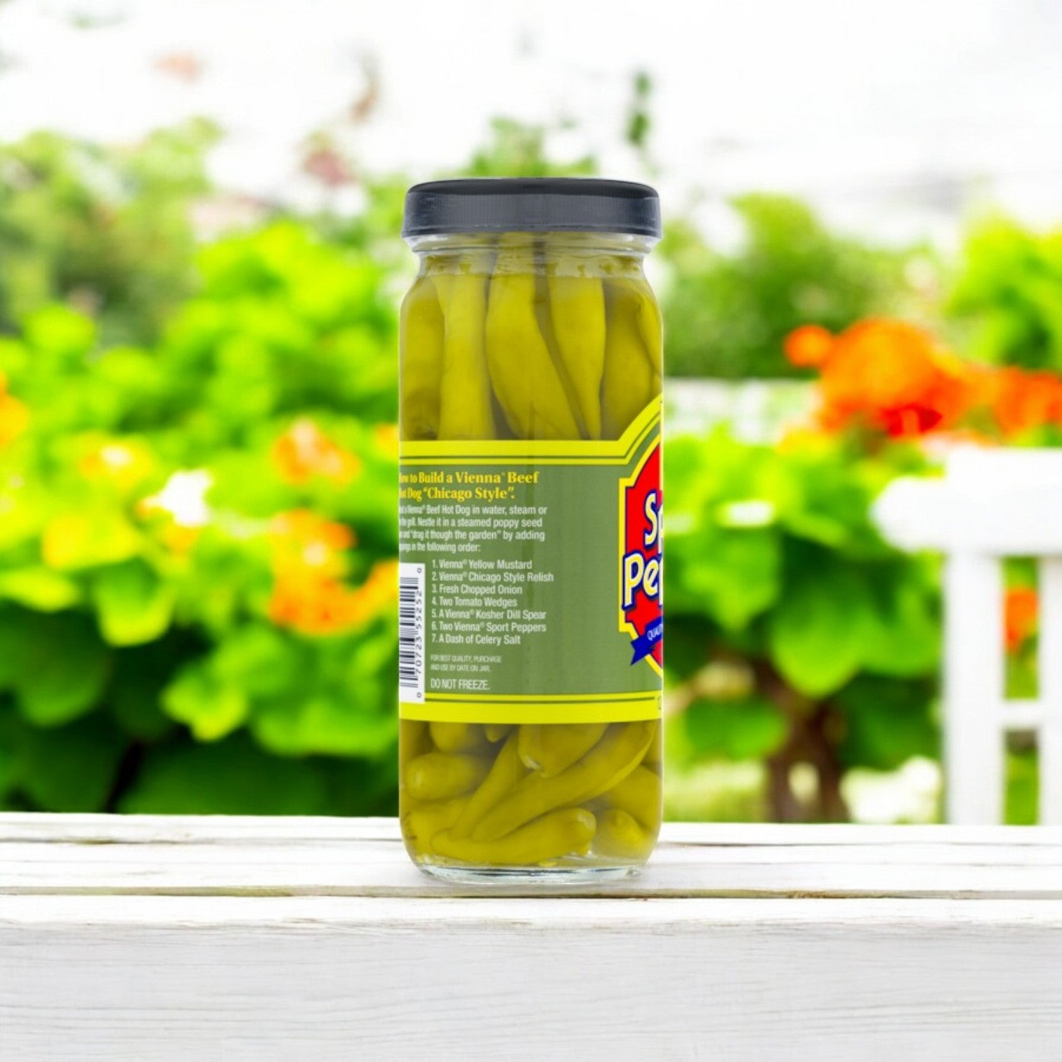 A 12 oz. jar of Vienna Beef Sport Peppers with a black lid rests on a wooden surface, surrounded by blurred green plants and bright orange flowers in the background.
