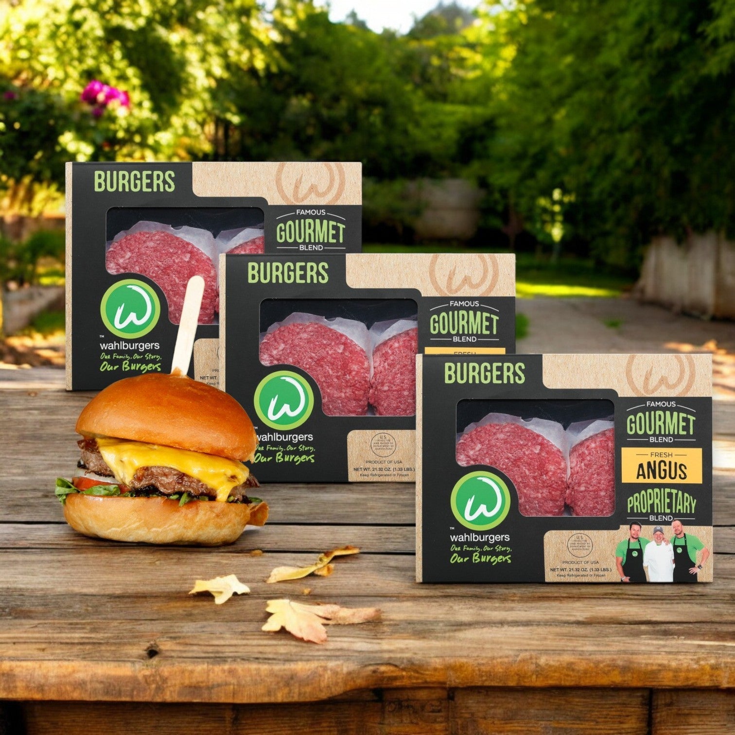 Three packages of Wahlburgers Famous Gourmet Blend Fresh Angus Burgers, 21.32 oz -3 Count, and one perfectly grilled burger are set on a wooden table outdoors against a lush leafy green backdrop.