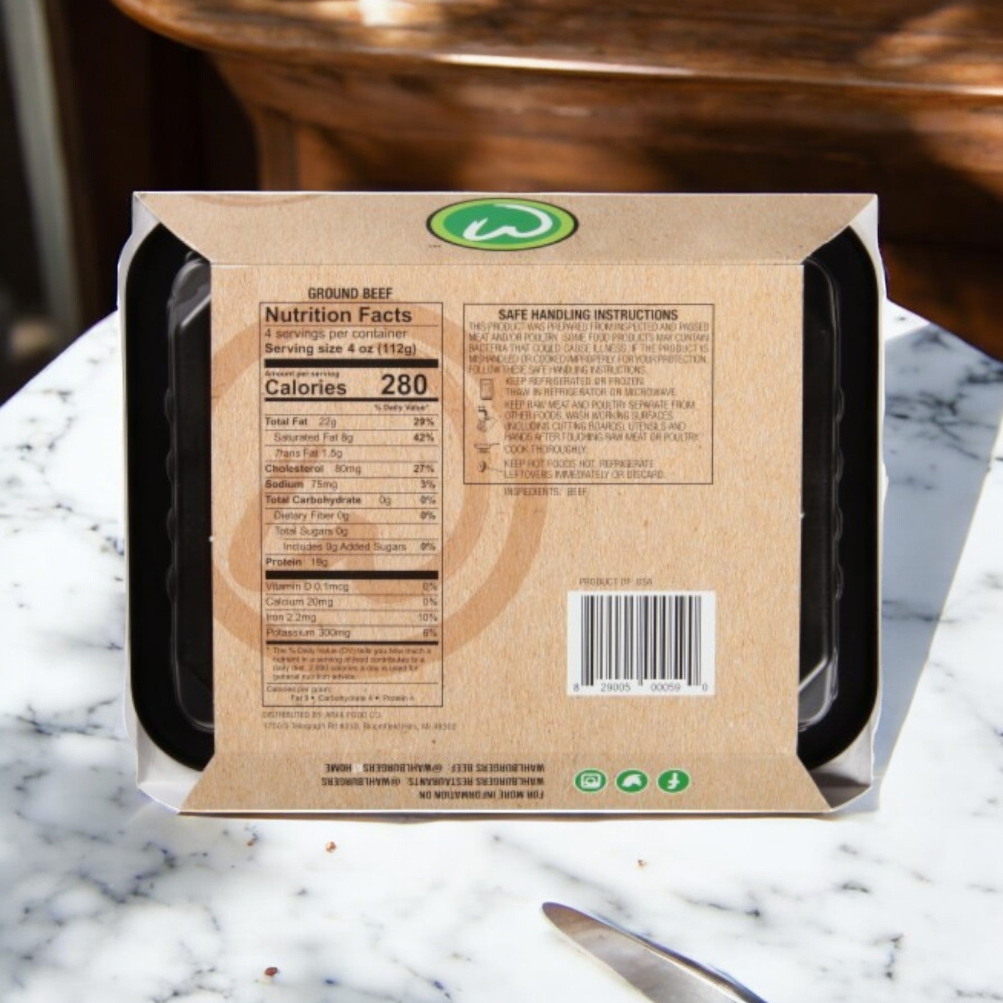 Brown cardboard packaging of Wahlburgers Signature Blend Loaf, 16 oz. - 3 Count on a marble table, showing nutrition facts and safe handling instructions with a knife visible in the corner.