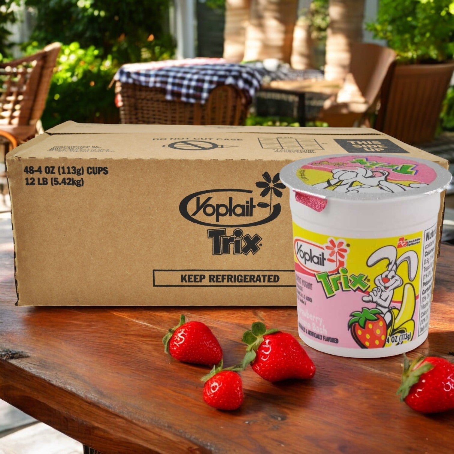 A box of Yoplait Trix Strawberry Banana Bash Yogurt, a popular kids snack, is on a wooden table beside one yogurt cup and three fresh strawberries.