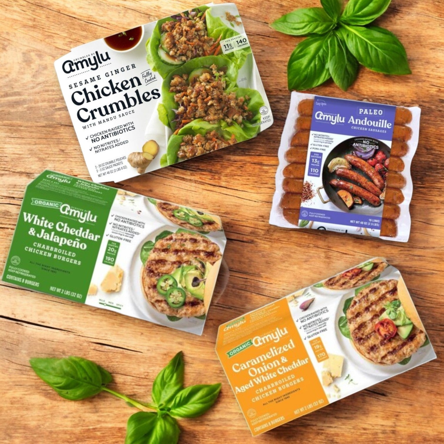 Four packaged foods rest on a wooden surface with basil leaves: Amylu’s 4-Pack Combo features Amylu Chicken Products like chicken crumbles, organic chicken burgers, white cheddar jalapeño options, and paleo-friendly andouille sausage caramelized onion burgers.