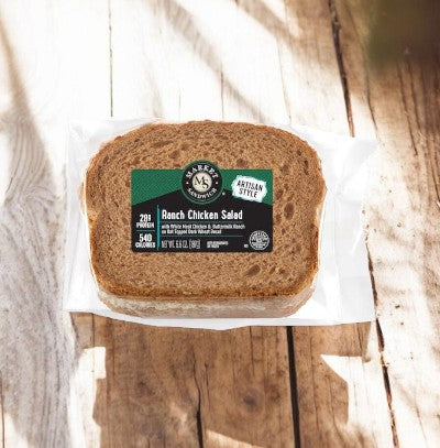 Under soft sunlight on a wooden surface, E.A. Sween's Ranch Chicken Salad Sandwich on multigrain bread ships frozen and boasts a 30-day shelf life in the cooler, promising to delight your taste buds.