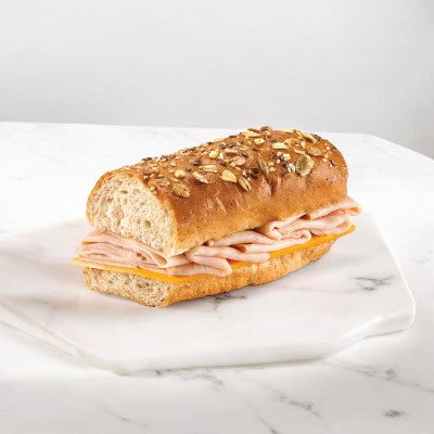 An E.A. Sween Smoked Turkey & Cheddar Sub Sandwich on a rustic oat-topped roll rests on a white marble surface.