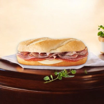 An E.A. Sween Italian Style Sub with premium cold cuts and cheese on a fresh sub roll rests on a white napkin atop wood. A small green plant adds a natural touch to this enticing frozen-to-cooler sandwich scene, enjoying a 30-day shelf life once thawed.