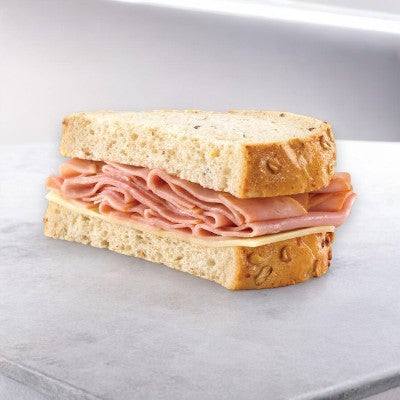 An E.A. Sween Ham & Swiss on rustic oat-topped wheat bread, featuring premium ham and cheese, rests elegantly on a light gray surface.